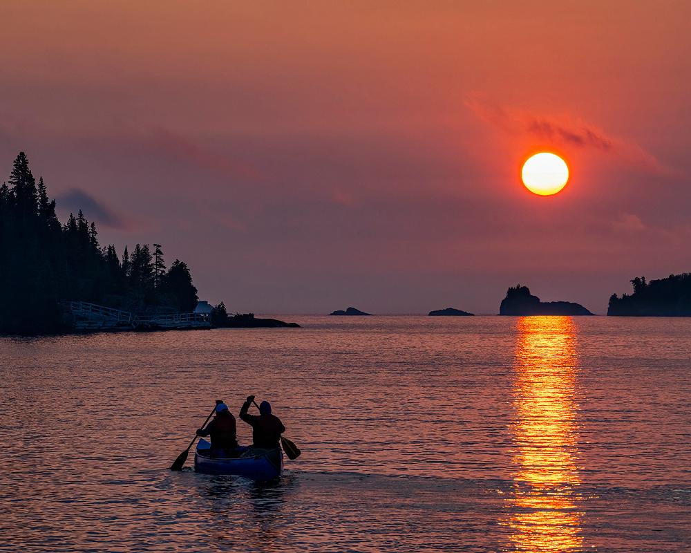 two people paddle a canoe in a bay as the sun rises over a series of islands